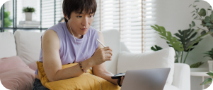 Person sitting on a couch while viewing laptop and holding mobile phone.